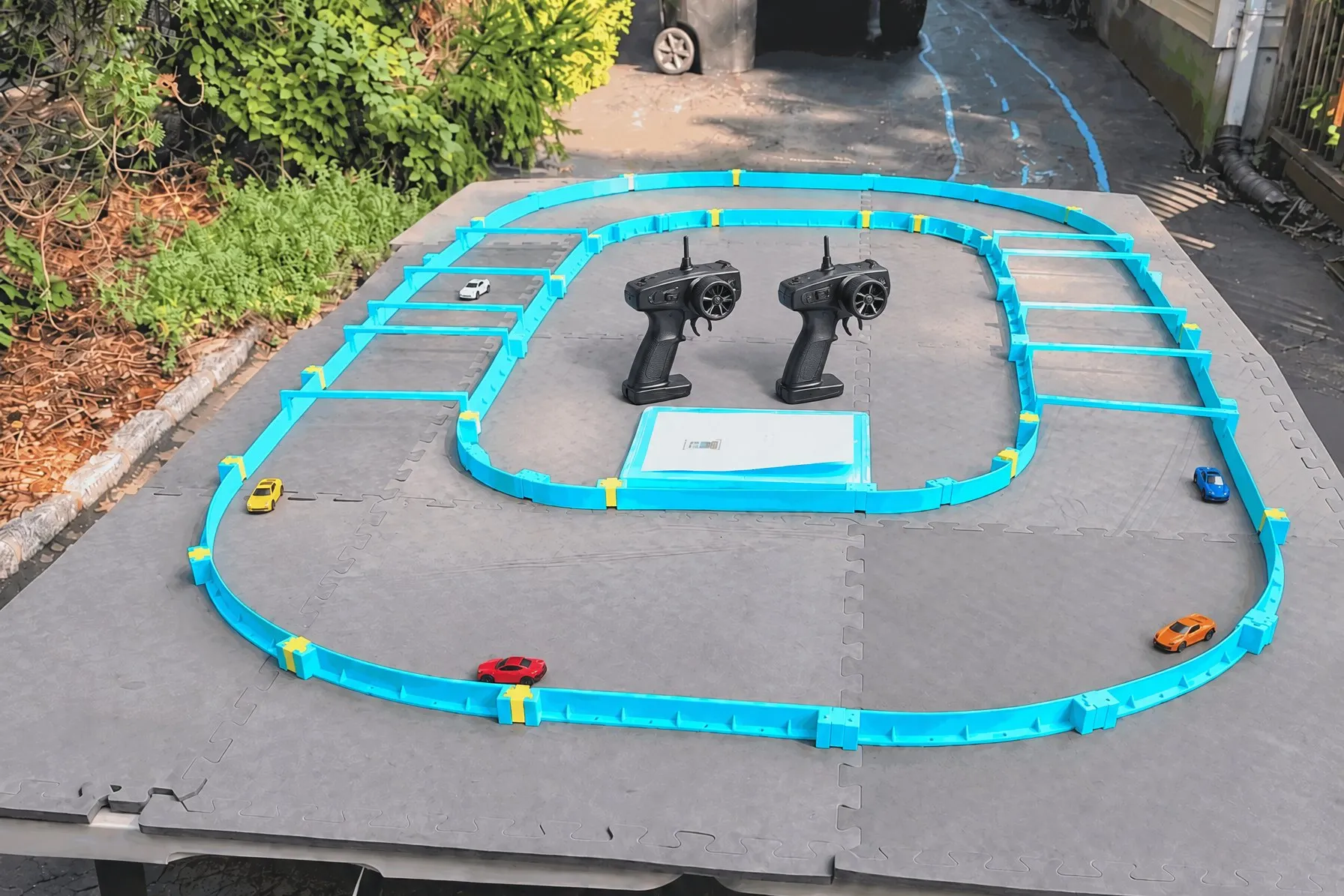 The Kit A race track set up on folding tables in a driveway: turquoise track walls on grey foam mats, five small micro RC cars spread around the oval, two pistol-grip controllers and a setup card in the infield.