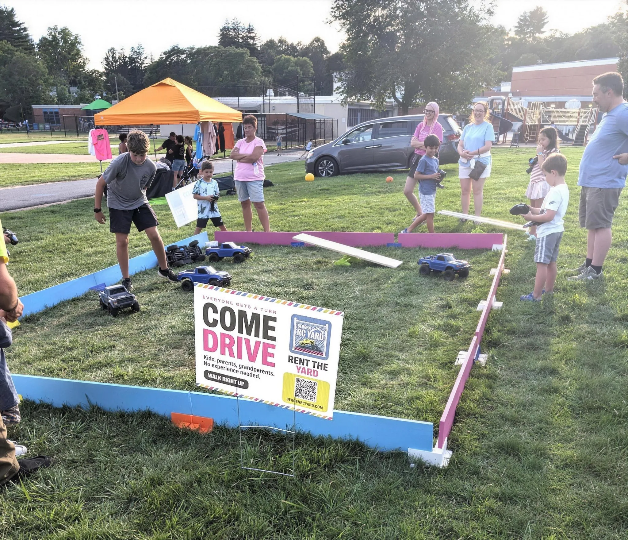 The drive yard at a summer event — 1/10-scale trucks and ramps inside blue and pink track walls on grass, drivers of all ages on the controllers, a Come Drive sign at the front wall.